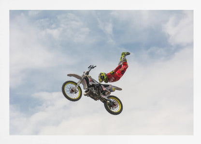 A motocross rider in red and yellow gear performs a high-flying stunt, arching their back while upside down in the air next to their dirt bike, set against a backdrop of a blue sky with white clouds and enclosed in a silver frame. Poster