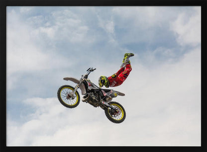 A motocross rider in red and yellow gear performs a high-flying stunt, arching their back while upside down in the air next to their dirt bike, set against a backdrop of a blue sky with white clouds and enclosed in a silver frame. Poster