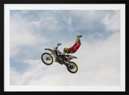 A motocross rider in red and yellow gear performs a high-flying stunt, arching their back while upside down in the air next to their dirt bike, set against a backdrop of a blue sky with white clouds and enclosed in a silver frame. Poster