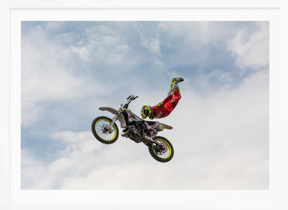 A motocross rider in red and yellow gear performs a high-flying stunt, arching their back while upside down in the air next to their dirt bike, set against a backdrop of a blue sky with white clouds and enclosed in a silver frame. Poster