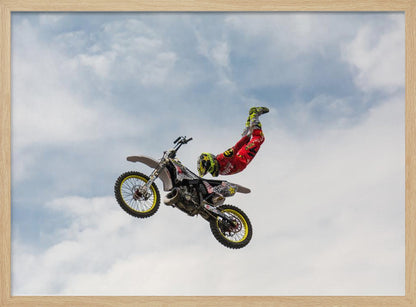 A motocross rider in red and yellow gear performs a high-flying stunt, arching their back while upside down in the air next to their dirt bike, set against a backdrop of a blue sky with white clouds and enclosed in a silver frame. Poster