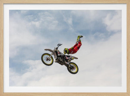 A motocross rider in red and yellow gear performs a high-flying stunt, arching their back while upside down in the air next to their dirt bike, set against a backdrop of a blue sky with white clouds and enclosed in a silver frame. Poster