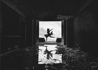 A dramatic black and white photo of a silhouetted dancer leaping in a bright doorway, viewed from inside a dark, derelict building. The dancer's reflection is captured in a puddle on the rubble-strewn floor. Poster