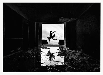 A dramatic black and white photo of a silhouetted dancer leaping in a bright doorway, viewed from inside a dark, derelict building. The dancer's reflection is captured in a puddle on the rubble-strewn floor. Poster