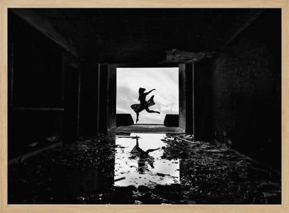 A dramatic black and white photo of a silhouetted dancer leaping in a bright doorway, viewed from inside a dark, derelict building. The dancer's reflection is captured in a puddle on the rubble-strewn floor. Poster