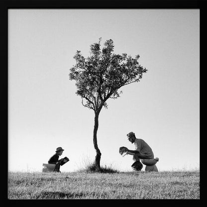 A framed, black and white photograph depicting a surreal scene of a man and a child sitting on toilets in an open grassy field under a single tree, both wearing hats and reading. Print