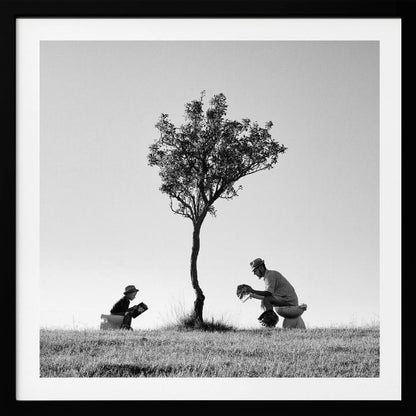 A framed, black and white photograph depicting a surreal scene of a man and a child sitting on toilets in an open grassy field under a single tree, both wearing hats and reading. Print