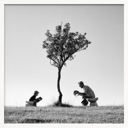 A framed, black and white photograph depicting a surreal scene of a man and a child sitting on toilets in an open grassy field under a single tree, both wearing hats and reading. Print