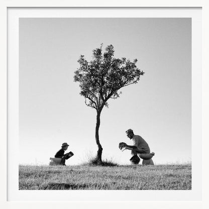 A framed, black and white photograph depicting a surreal scene of a man and a child sitting on toilets in an open grassy field under a single tree, both wearing hats and reading. Print