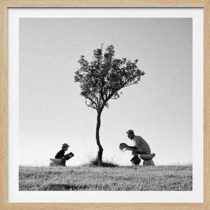 A framed, black and white photograph depicting a surreal scene of a man and a child sitting on toilets in an open grassy field under a single tree, both wearing hats and reading. Print