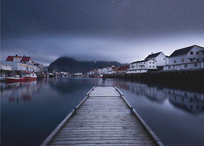 A long exposure photograph of a serene coastal town at night, viewed from a wooden pier that leads into calm, dark water. A lone person stands at the end of the pier, gazing at the town's reflected lights. Behind the village, a mountain looms under a breathtaking, star-filled sky. The image is presented within a silver-colored frame. Artwork
