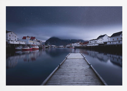 A long exposure photograph of a serene coastal town at night, viewed from a wooden pier that leads into calm, dark water. A lone person stands at the end of the pier, gazing at the town's reflected lights. Behind the village, a mountain looms under a breathtaking, star-filled sky. The image is presented within a silver-colored frame. Artwork