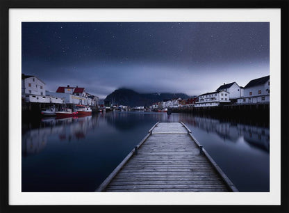 A long exposure photograph of a serene coastal town at night, viewed from a wooden pier that leads into calm, dark water. A lone person stands at the end of the pier, gazing at the town's reflected lights. Behind the village, a mountain looms under a breathtaking, star-filled sky. The image is presented within a silver-colored frame. Artwork