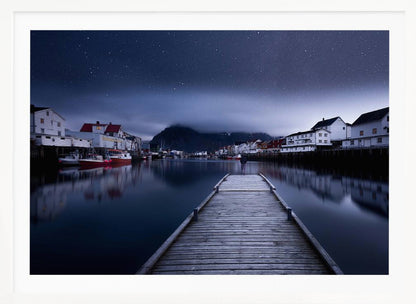 A long exposure photograph of a serene coastal town at night, viewed from a wooden pier that leads into calm, dark water. A lone person stands at the end of the pier, gazing at the town's reflected lights. Behind the village, a mountain looms under a breathtaking, star-filled sky. The image is presented within a silver-colored frame. Artwork