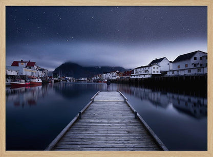 A long exposure photograph of a serene coastal town at night, viewed from a wooden pier that leads into calm, dark water. A lone person stands at the end of the pier, gazing at the town's reflected lights. Behind the village, a mountain looms under a breathtaking, star-filled sky. The image is presented within a silver-colored frame. Artwork
