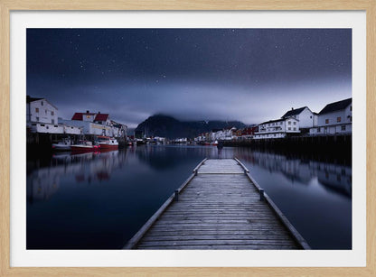 A long exposure photograph of a serene coastal town at night, viewed from a wooden pier that leads into calm, dark water. A lone person stands at the end of the pier, gazing at the town's reflected lights. Behind the village, a mountain looms under a breathtaking, star-filled sky. The image is presented within a silver-colored frame. Artwork