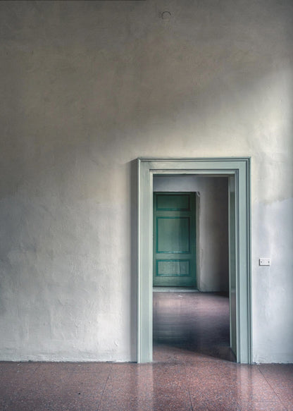 A minimalist photograph of a room with grey, textured walls and a reddish-brown floor, focusing on an open, light green doorway that reveals another room with a closed, darker green door, creating a sense of depth and perspective. Wall Art