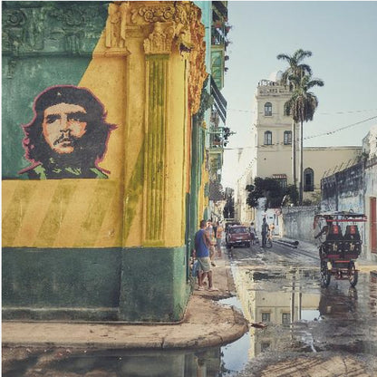 A framed photograph of a vibrant street corner in a city like Havana, Cuba, with a large mural of Che Guevara on a yellow and green wall. The wet street reflects the buildings and a palm tree, while people, a vintage car, and a rickshaw occupy the narrow road. Poster