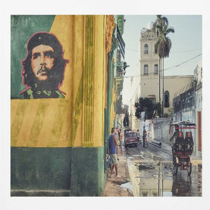 A framed photograph of a vibrant street corner in a city like Havana, Cuba, with a large mural of Che Guevara on a yellow and green wall. The wet street reflects the buildings and a palm tree, while people, a vintage car, and a rickshaw occupy the narrow road. Poster