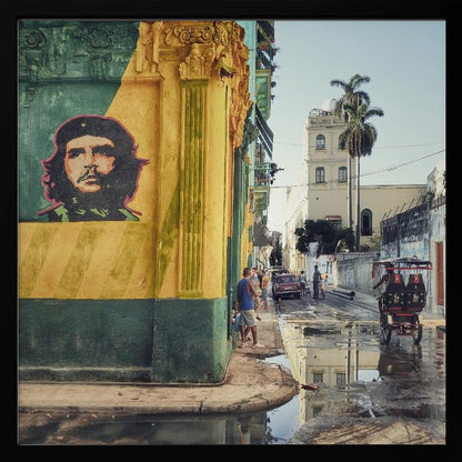 A framed photograph of a vibrant street corner in a city like Havana, Cuba, with a large mural of Che Guevara on a yellow and green wall. The wet street reflects the buildings and a palm tree, while people, a vintage car, and a rickshaw occupy the narrow road. Poster