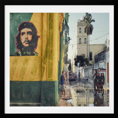A framed photograph of a vibrant street corner in a city like Havana, Cuba, with a large mural of Che Guevara on a yellow and green wall. The wet street reflects the buildings and a palm tree, while people, a vintage car, and a rickshaw occupy the narrow road. Poster