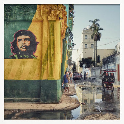 A framed photograph of a vibrant street corner in a city like Havana, Cuba, with a large mural of Che Guevara on a yellow and green wall. The wet street reflects the buildings and a palm tree, while people, a vintage car, and a rickshaw occupy the narrow road. Poster