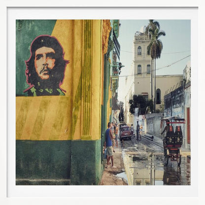 A framed photograph of a vibrant street corner in a city like Havana, Cuba, with a large mural of Che Guevara on a yellow and green wall. The wet street reflects the buildings and a palm tree, while people, a vintage car, and a rickshaw occupy the narrow road. Poster