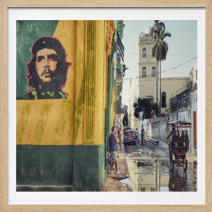 A framed photograph of a vibrant street corner in a city like Havana, Cuba, with a large mural of Che Guevara on a yellow and green wall. The wet street reflects the buildings and a palm tree, while people, a vintage car, and a rickshaw occupy the narrow road. Poster