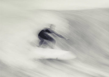 An artistic black and white photograph using motion blur to capture a surfer in a dynamic, fluid pose on a wave. The slow shutter speed creates a sense of speed and abstract movement, with the surfer's dark wetsuit contrasting against the white, streaked water. The image is presented in a silver frame. Decor