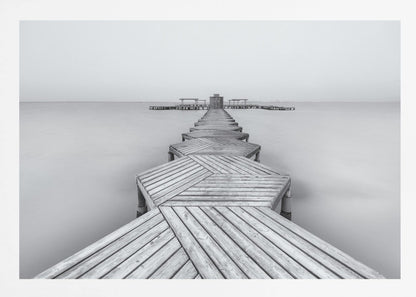 A framed, black and white photograph capturing a long wooden pier from a first-person perspective. The pier, constructed from a series of hexagonal platforms, stretches out into a calm, placid body of water that disappears into a foggy horizon, creating a serene and minimalist scene. Print