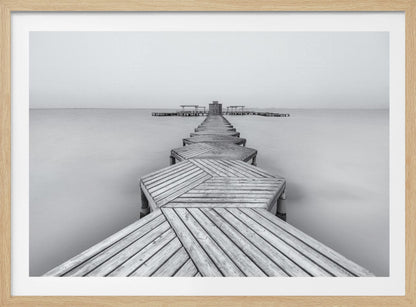 A framed, black and white photograph capturing a long wooden pier from a first-person perspective. The pier, constructed from a series of hexagonal platforms, stretches out into a calm, placid body of water that disappears into a foggy horizon, creating a serene and minimalist scene. Print