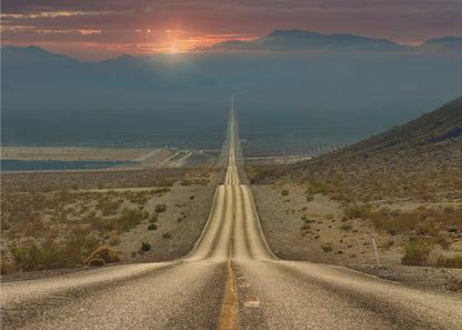A high-angle view of a long, straight, and hilly road disappearing into the horizon of a vast desert valley at sunset, with mountains in the background under a warm, glowing sky, all enclosed in a silver frame. Poster
