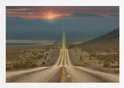 A high-angle view of a long, straight, and hilly road disappearing into the horizon of a vast desert valley at sunset, with mountains in the background under a warm, glowing sky, all enclosed in a silver frame. Poster