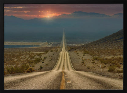 A high-angle view of a long, straight, and hilly road disappearing into the horizon of a vast desert valley at sunset, with mountains in the background under a warm, glowing sky, all enclosed in a silver frame. Poster