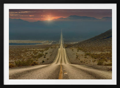 A high-angle view of a long, straight, and hilly road disappearing into the horizon of a vast desert valley at sunset, with mountains in the background under a warm, glowing sky, all enclosed in a silver frame. Poster