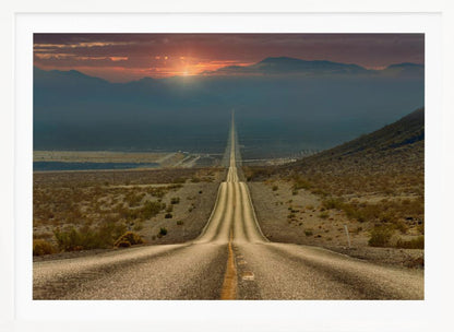 A high-angle view of a long, straight, and hilly road disappearing into the horizon of a vast desert valley at sunset, with mountains in the background under a warm, glowing sky, all enclosed in a silver frame. Poster