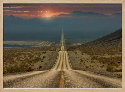 A high-angle view of a long, straight, and hilly road disappearing into the horizon of a vast desert valley at sunset, with mountains in the background under a warm, glowing sky, all enclosed in a silver frame. Poster