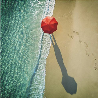An aerial photograph of a serene beach scene, featuring a vibrant red umbrella on the golden sand next to the clear turquoise ocean. Gentle white waves lap the shore, and a long shadow from the umbrella stretches across the sand, where a trail of footprints leads away. Poster
