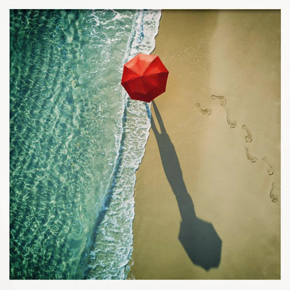 An aerial photograph of a serene beach scene, featuring a vibrant red umbrella on the golden sand next to the clear turquoise ocean. Gentle white waves lap the shore, and a long shadow from the umbrella stretches across the sand, where a trail of footprints leads away. Poster