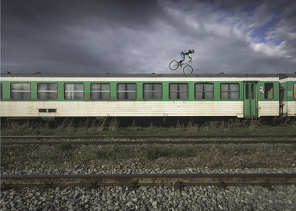 A BMX rider is captured in mid-air performing a stunt on the roof of a green and white passenger train car, set against a dramatic, stormy sky. The scene takes place on railway tracks with gravel and overgrown grass, and the entire image is enclosed in a silver frame. Decor