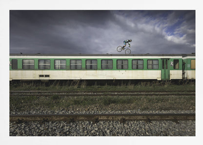 A BMX rider is captured in mid-air performing a stunt on the roof of a green and white passenger train car, set against a dramatic, stormy sky. The scene takes place on railway tracks with gravel and overgrown grass, and the entire image is enclosed in a silver frame. Decor