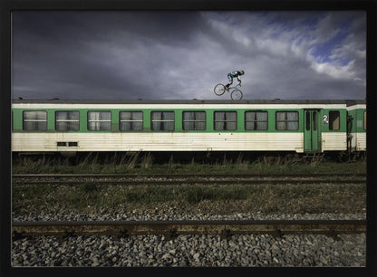 A BMX rider is captured in mid-air performing a stunt on the roof of a green and white passenger train car, set against a dramatic, stormy sky. The scene takes place on railway tracks with gravel and overgrown grass, and the entire image is enclosed in a silver frame. Decor