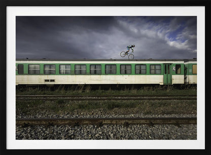 A BMX rider is captured in mid-air performing a stunt on the roof of a green and white passenger train car, set against a dramatic, stormy sky. The scene takes place on railway tracks with gravel and overgrown grass, and the entire image is enclosed in a silver frame. Decor