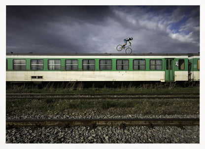 A BMX rider is captured in mid-air performing a stunt on the roof of a green and white passenger train car, set against a dramatic, stormy sky. The scene takes place on railway tracks with gravel and overgrown grass, and the entire image is enclosed in a silver frame. Decor