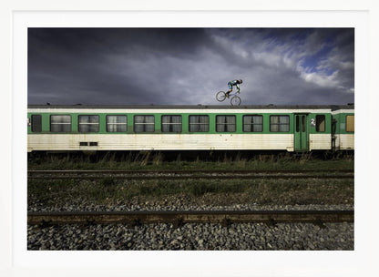 A BMX rider is captured in mid-air performing a stunt on the roof of a green and white passenger train car, set against a dramatic, stormy sky. The scene takes place on railway tracks with gravel and overgrown grass, and the entire image is enclosed in a silver frame. Decor