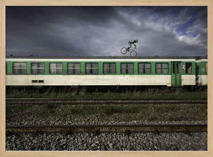 A BMX rider is captured in mid-air performing a stunt on the roof of a green and white passenger train car, set against a dramatic, stormy sky. The scene takes place on railway tracks with gravel and overgrown grass, and the entire image is enclosed in a silver frame. Decor