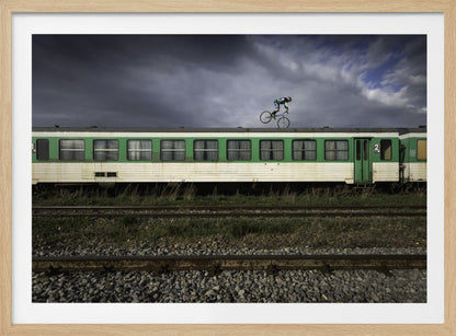 A BMX rider is captured in mid-air performing a stunt on the roof of a green and white passenger train car, set against a dramatic, stormy sky. The scene takes place on railway tracks with gravel and overgrown grass, and the entire image is enclosed in a silver frame. Decor