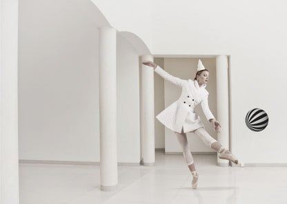 A ballerina dressed in a white quilted coat and a pointed hat dances en pointe in a minimalist white hall with large columns. She gracefully extends her arms, looking towards a black and white striped ball that appears to be floating in mid-air. The scene is presented within a silver picture frame. Wall Art