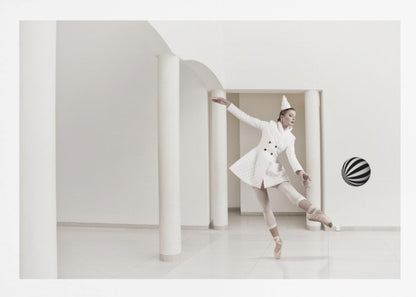 A ballerina dressed in a white quilted coat and a pointed hat dances en pointe in a minimalist white hall with large columns. She gracefully extends her arms, looking towards a black and white striped ball that appears to be floating in mid-air. The scene is presented within a silver picture frame. Wall Art