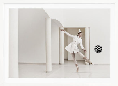 A ballerina dressed in a white quilted coat and a pointed hat dances en pointe in a minimalist white hall with large columns. She gracefully extends her arms, looking towards a black and white striped ball that appears to be floating in mid-air. The scene is presented within a silver picture frame. Wall Art